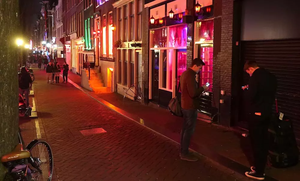 twee mannen die hun smartphone vasthouden en buiten een gebouw in de Wallen, Amsterdam staan, verlicht door nachtlampjes.
