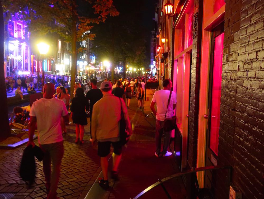 tourists walking on the Oudezijds Achtburgwal that is also known as the famous red street in Amsterdam with red light windows on the right side of the street.