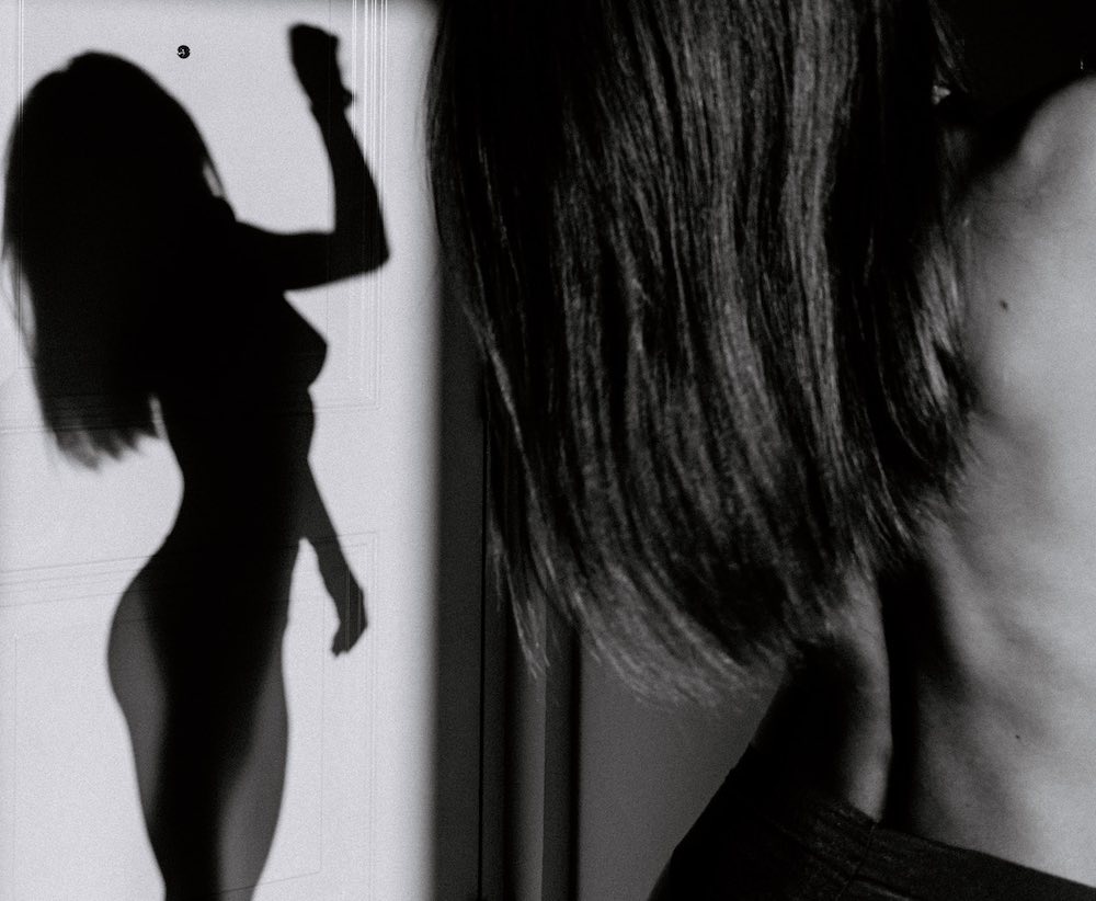 Grayscale image showing a sex worker's profile and shoulder-length hair on the right; on the left, their shadow is cast on a wall in a dramatic silhouette.