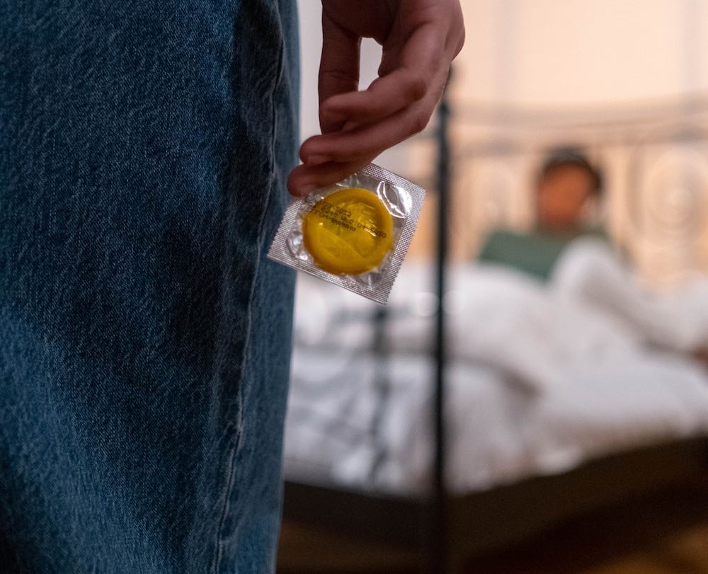 A man with a condom in his hand walking towards a bed, while another gay men is waiting