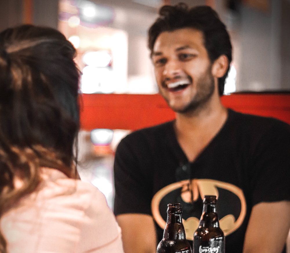 young man in black Batman t-shirt laughing while looking at his female date in a bar