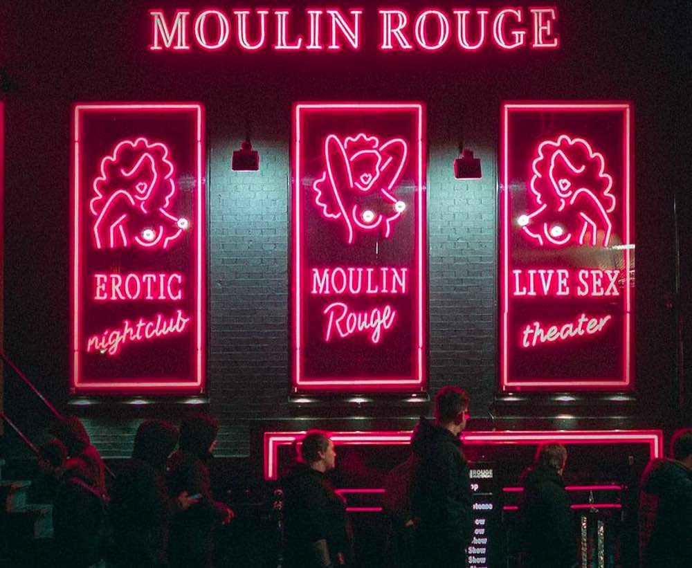 the frontview of Moulin Rouge in Amsterdam Red Light District at night with red-lit neon and people in front of the building