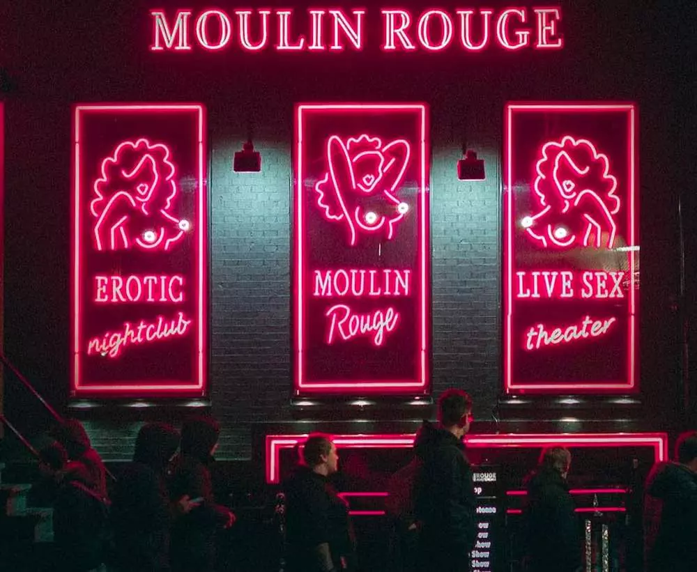 Die Vorderansicht des Moulin Rouge im Amsterdamer Rotlichtviertel bei Nacht mit rot beleuchteten Neonreklamen und Menschen vor dem Gebäude