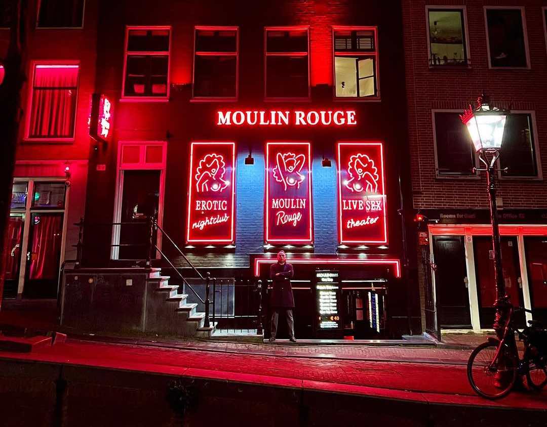 The front view of Moulin Rouge in De Wallen in Amsterdam at night, showing the red neon logo and a bouncer in front of the door.