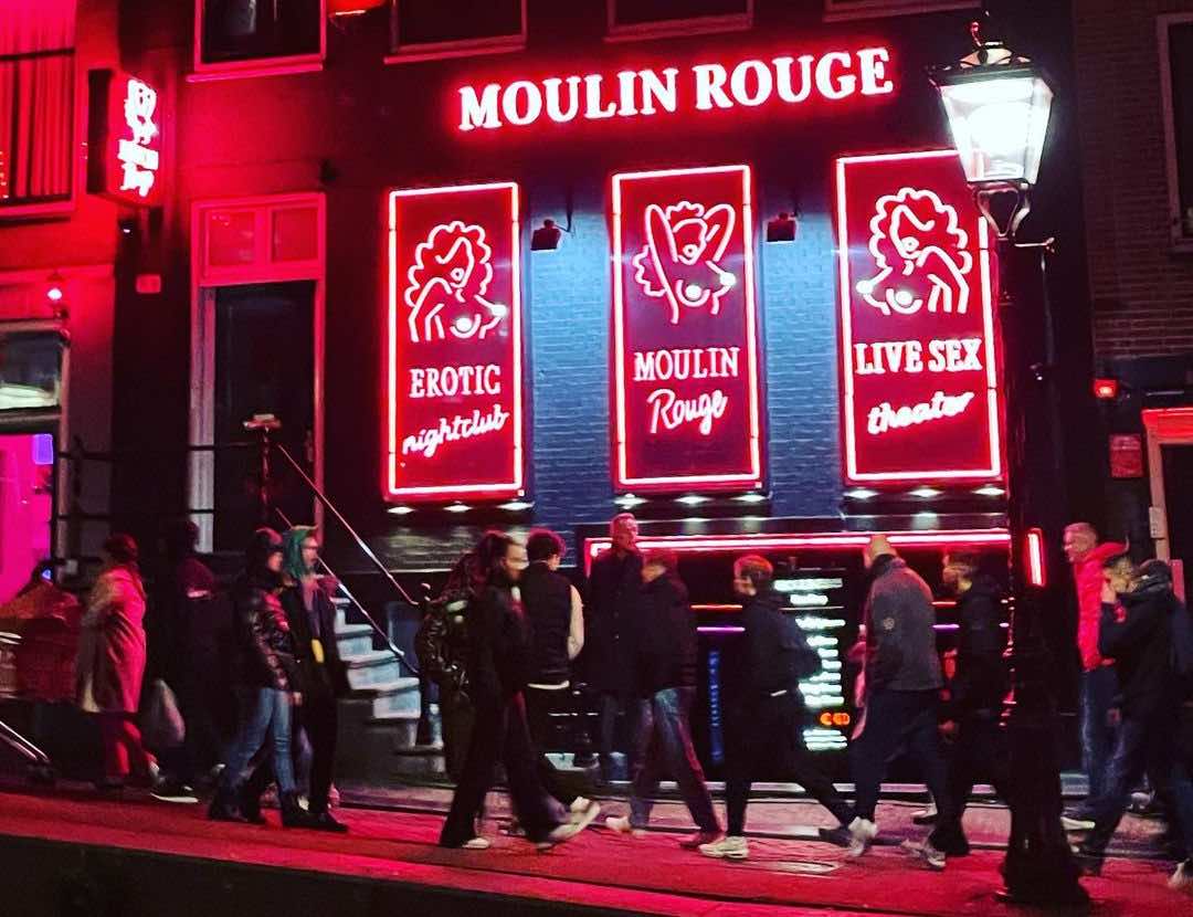 people walking in Amsterdam Red Light District with Moulin Rouge on the right side with red-lit neon advertisements 