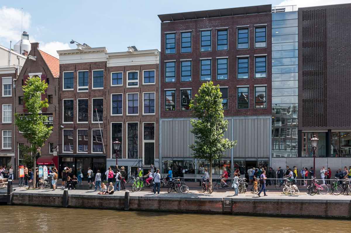 The front of the Anne Frank House in Amsterdam with a canal and street with people in front of it taken on a sunny, summer day