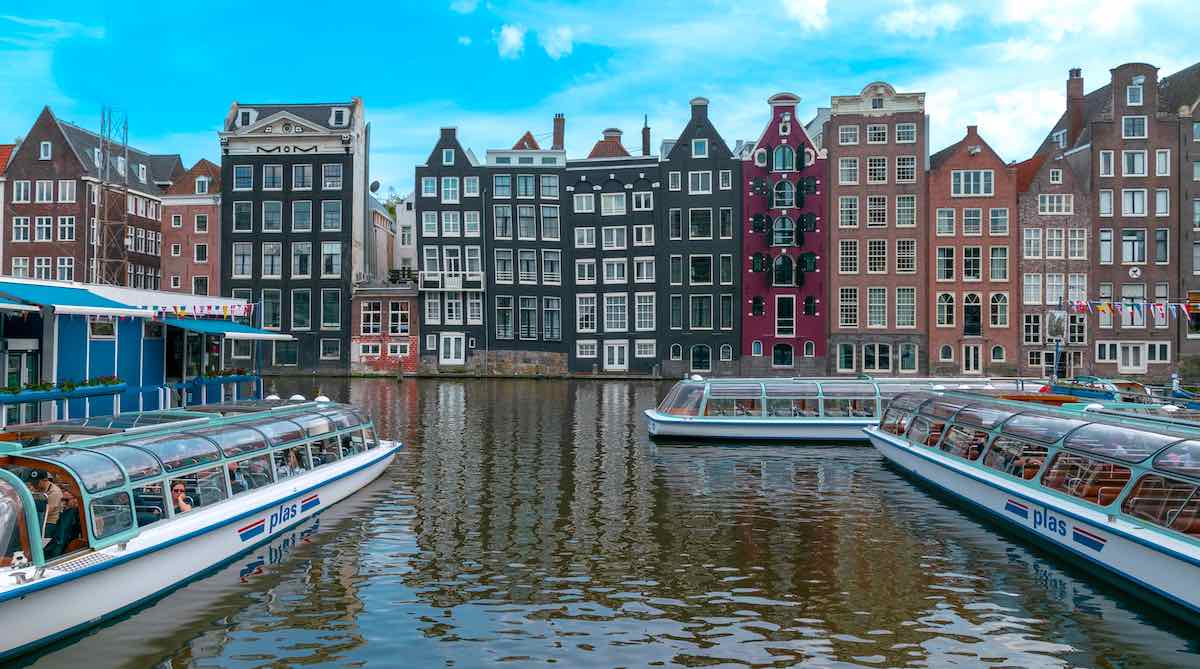 3 white canal boats in Amsterdam in front several canal houses with blue sky