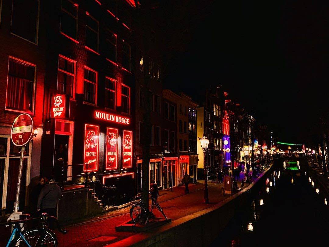 A night shot of De Wallen in Amsterdam showing red-lit neons on the left side and a canal in the middle.