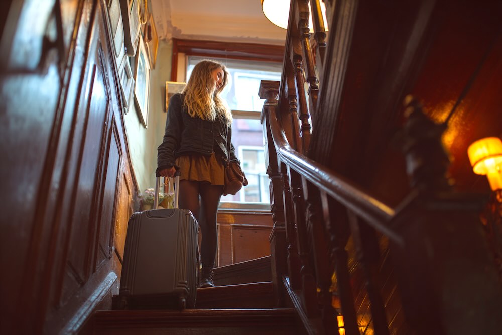 Attractive Female Tourist with a small, modern suitcase climbing steep narrow stairs inside a historic Amsterdam home, wooden interior, tight space, warm lighting, authentic Dutch atmosphere