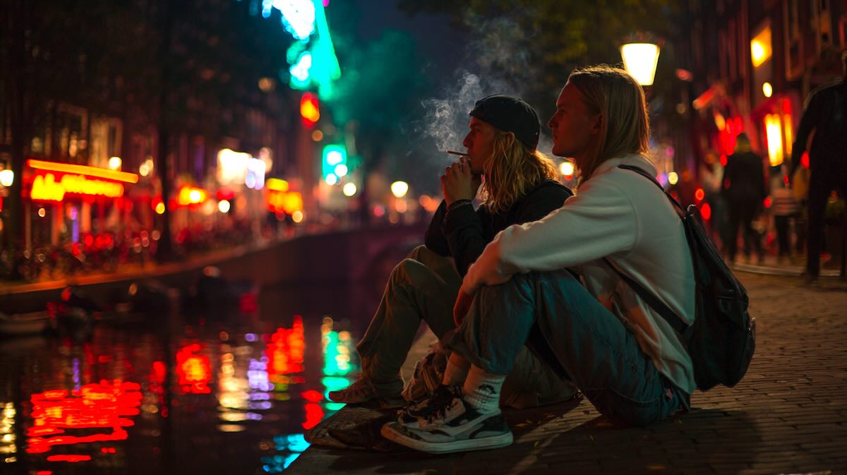 Two young tourists sitting by a canal in Amsterdam’s Red Light District at night, subtle neon reflections on the water, casually smoking weed, relaxed and safe atmosphere, 